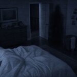 Toby Atividade Paranormal - Quarto escuro visto por uma câmera noturna, com uma cama em primeiro plano, uma porta entreaberta ao fundo e uma forma humanoide quase invisível misturada às sombras próximas ao corredor, inspirada na presença de Toby em Atividade Paranormal.