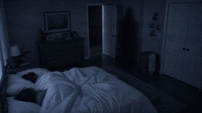 Toby Atividade Paranormal - Quarto escuro visto por uma câmera noturna, com uma cama em primeiro plano, uma porta entreaberta ao fundo e uma forma humanoide quase invisível misturada às sombras próximas ao corredor, inspirada na presença de Toby em Atividade Paranormal.