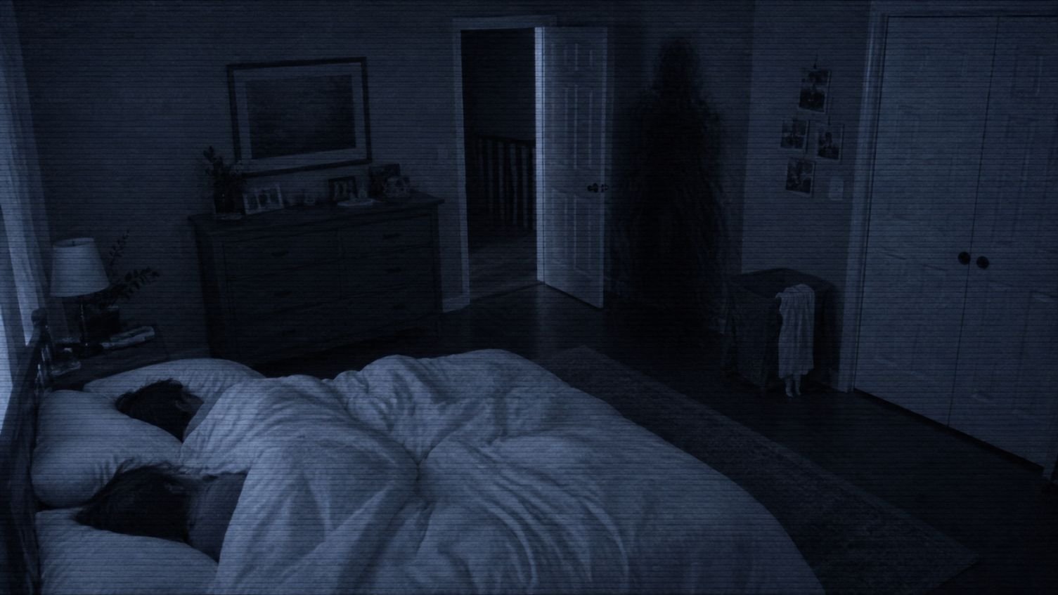 Toby Atividade Paranormal - Quarto escuro visto por uma câmera noturna, com uma cama em primeiro plano, uma porta entreaberta ao fundo e uma forma humanoide quase invisível misturada às sombras próximas ao corredor, inspirada na presença de Toby em Atividade Paranormal.